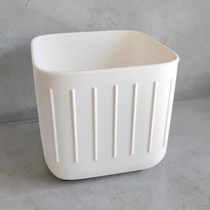 May include: A white, square-shaped storage container with vertical, ridged detailing. The container has rounded corners and a smooth, matte finish. The design is simple and modern, suitable for various organizational needs.