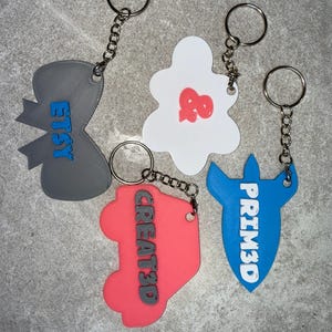 May include: A collection of keychains in various shapes and colors. One is gray with "ETSY" in blue, another is white with a red design, a third is coral with "CREATED" in black, and the last is blue with "PRIM3D" in white.