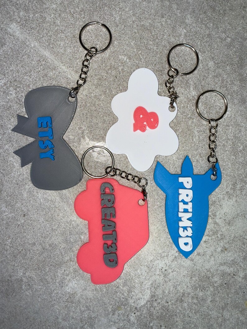 May include: A collection of keychains in various shapes and colors. One is gray with "ETSY" in blue, another is white with a red design, a third is coral with "CREATED" in black, and the last is blue with "PRIM3D" in white.