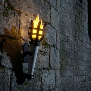 May include: A wall-mounted torch with a bright yellow flame, set against a stone wall. The torch has a black metal cage around the flame and a white cylindrical base. The scene is lit with a warm glow.