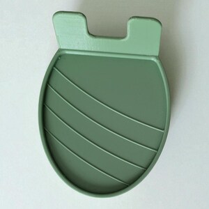 May include: A sage green, oval-shaped coaster with a raised, ridged design and two small, rectangular tabs at the top. The coaster is made of a solid, matte material and is designed to protect surfaces from heat and moisture.