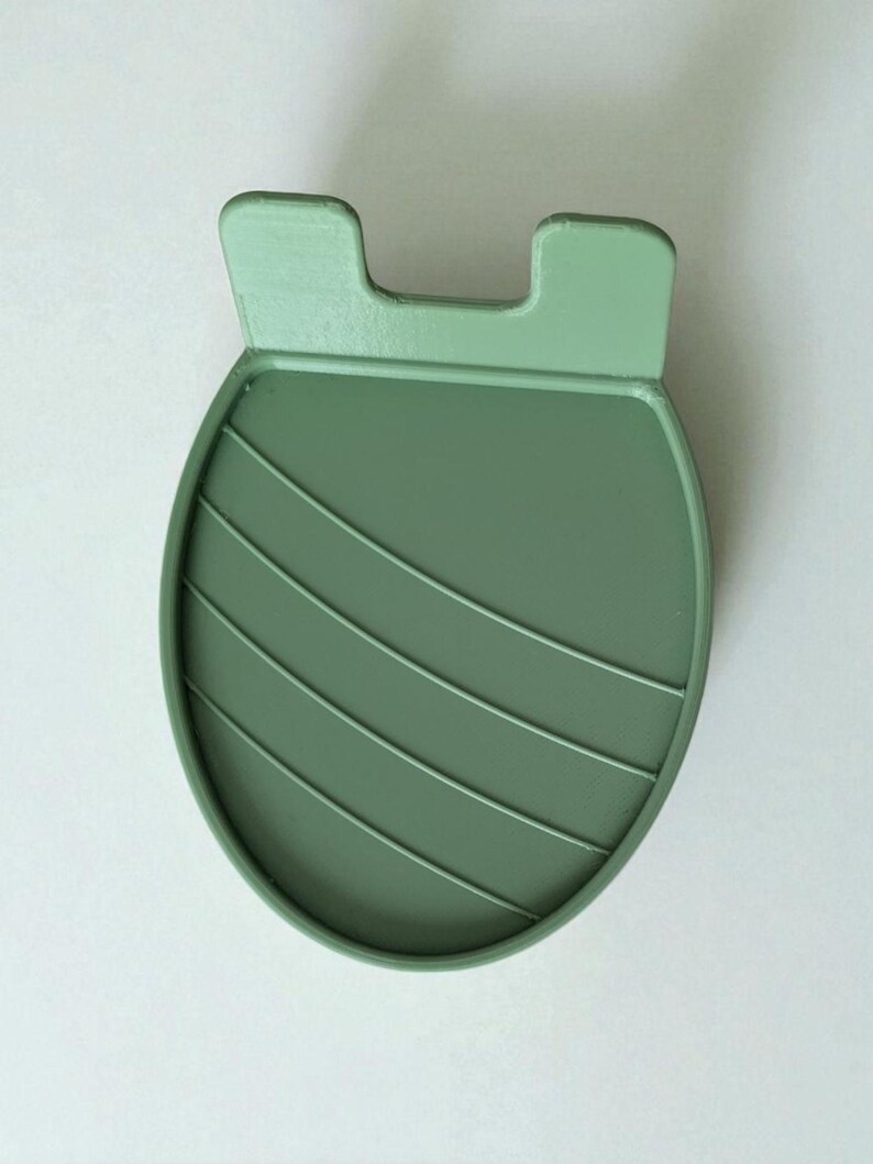May include: A sage green, oval-shaped coaster with a raised, ridged design and two small, rectangular tabs at the top. The coaster is made of a solid, matte material and is designed to protect surfaces from heat and moisture.