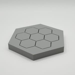May include: A grey hexagonal coaster with a honeycomb pattern. The coaster has seven smaller hexagonal shapes outlined in black, set within the larger hexagon. The coaster is made of a solid, matte material.