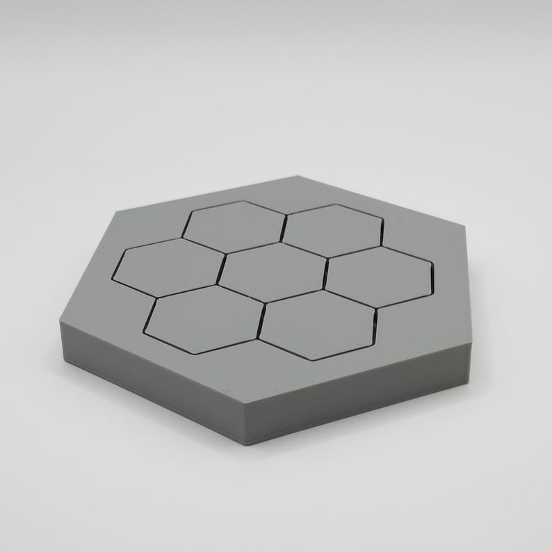 May include: A grey hexagonal coaster with a honeycomb pattern. The coaster has seven smaller hexagonal shapes outlined in black, set within the larger hexagon. The coaster is made of a solid, matte material.