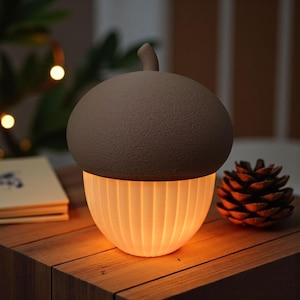 May include: An acorn-shaped night light with a brown top and a glowing, ribbed white base. The lamp sits on a wooden surface, with a pine cone and a book in the background. The lamp emits a warm, inviting light.