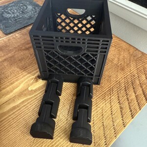 May include: A black plastic crate with a diamond pattern, set on black articulated legs. The crate has a rectangular shape with a handle cutout on the front. Inside the crate is a smaller, diamond-patterned container.
