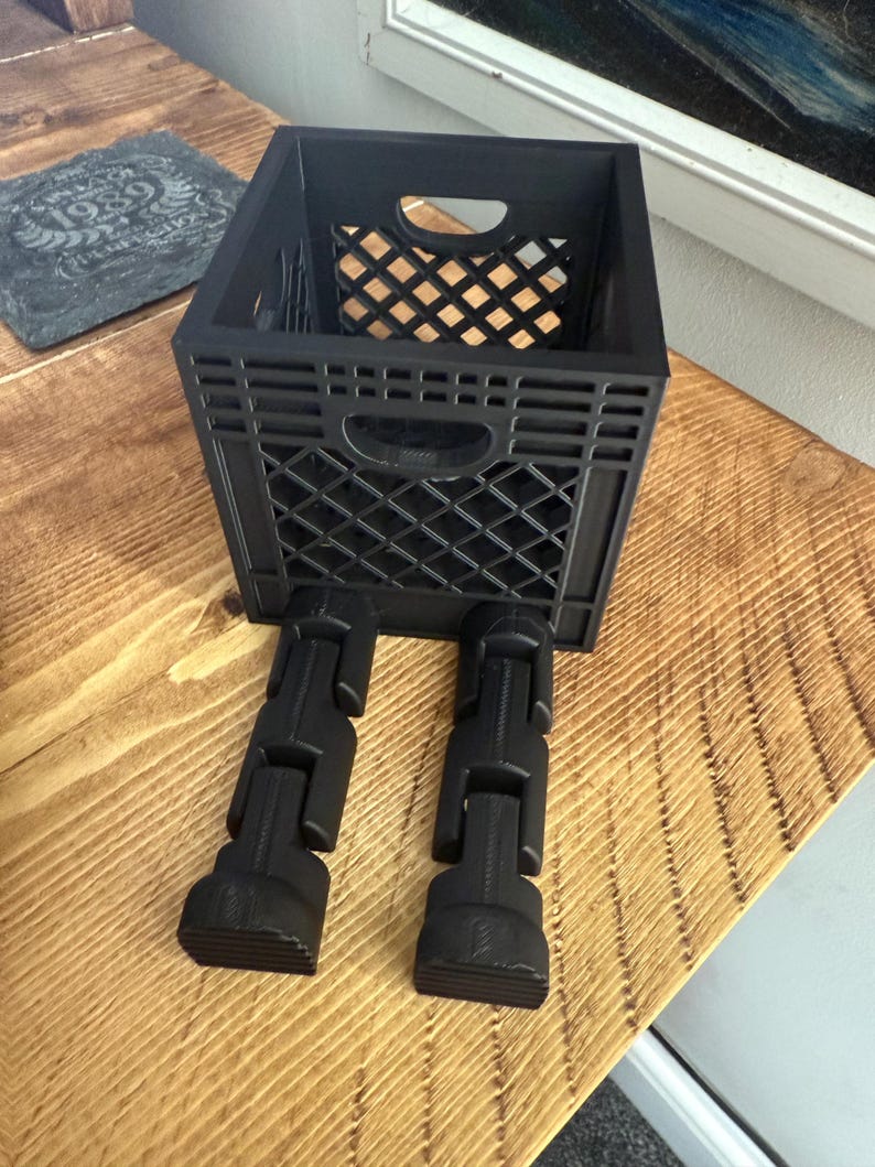May include: A black plastic crate with a diamond pattern, set on black articulated legs. The crate has a rectangular shape with a handle cutout on the front. Inside the crate is a smaller, diamond-patterned container.