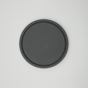 May include: A round, dark gray coaster with a raised, black rim. The coaster is made of a solid material and is designed to protect surfaces from drink condensation. The simple design is suitable for various home decor styles.