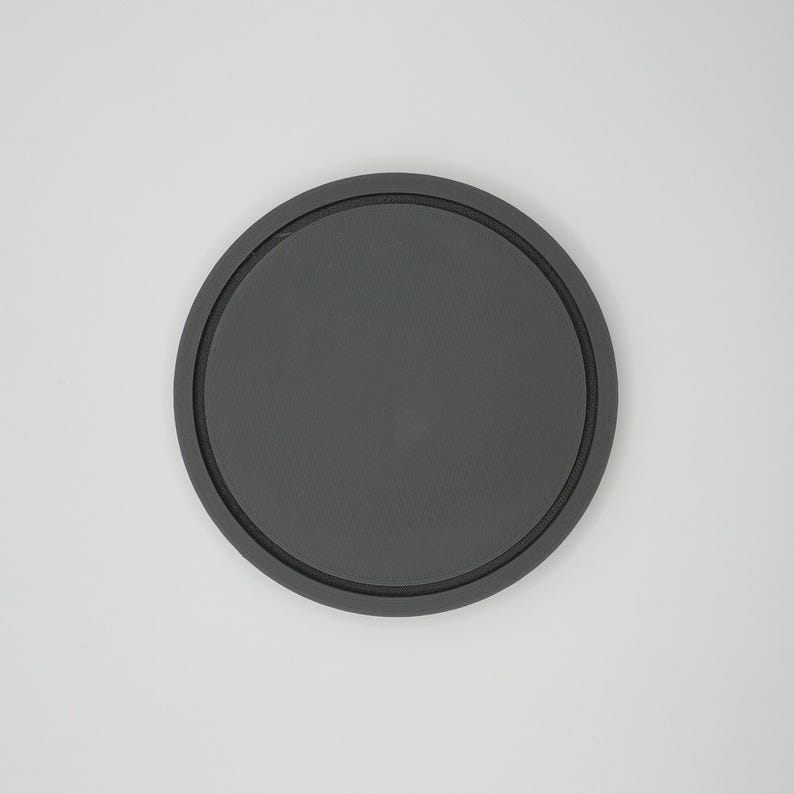 May include: A round, dark gray coaster with a raised, black rim. The coaster is made of a solid material and is designed to protect surfaces from drink condensation. The simple design is suitable for various home decor styles.