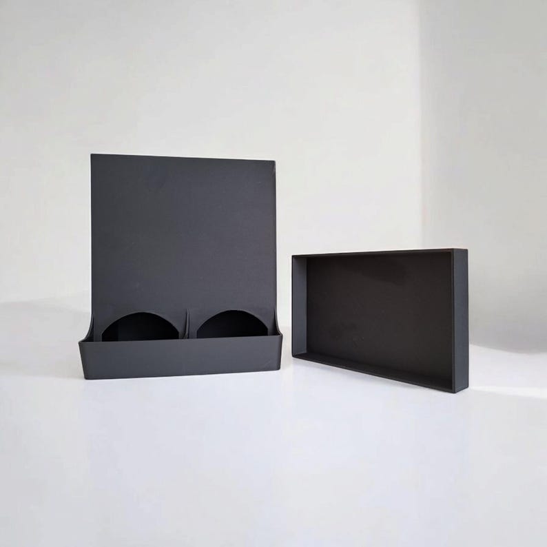 May include: Two-piece black display set. The set includes a tall, rectangular stand with two curved cutouts and a separate, rectangular tray. The items are made of a matte black material and are set against a white background.