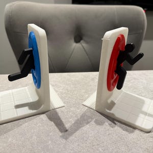 May include: A pair of white bookends with a portal design. One bookend has a blue portal with a black figure, the other has a red portal with a black figure. The bookends are on a white surface.