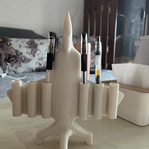 May include: A cream-colored pen holder shaped like an airplane. The desk organizer has multiple cylindrical slots to hold pens and pencils. Several pens are visible in the slots. The holder has a three-legged base and a pointed nose.