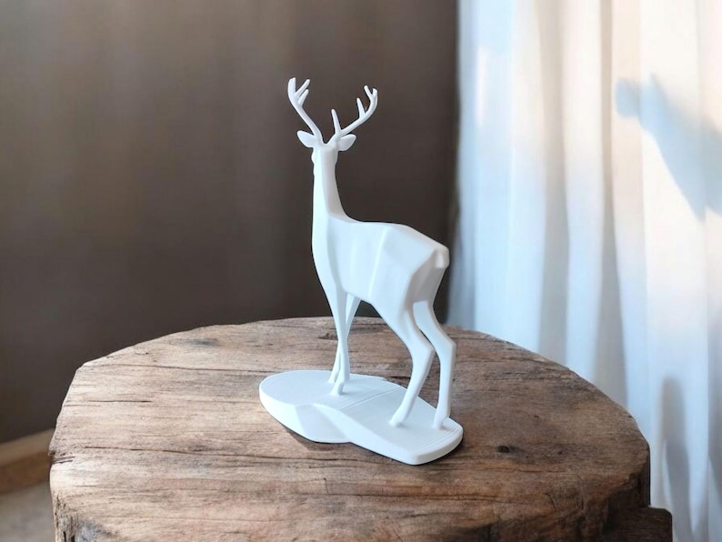Puede incluir: Una escultura de ciervo geom&eacute;trico blanco con cuernos se alza sobre una base blanca. El ciervo est&aacute; colocado sobre una mesa de madera redonda y r&uacute;stica. El fondo presenta una pared marr&oacute;n y una cortina blanca.