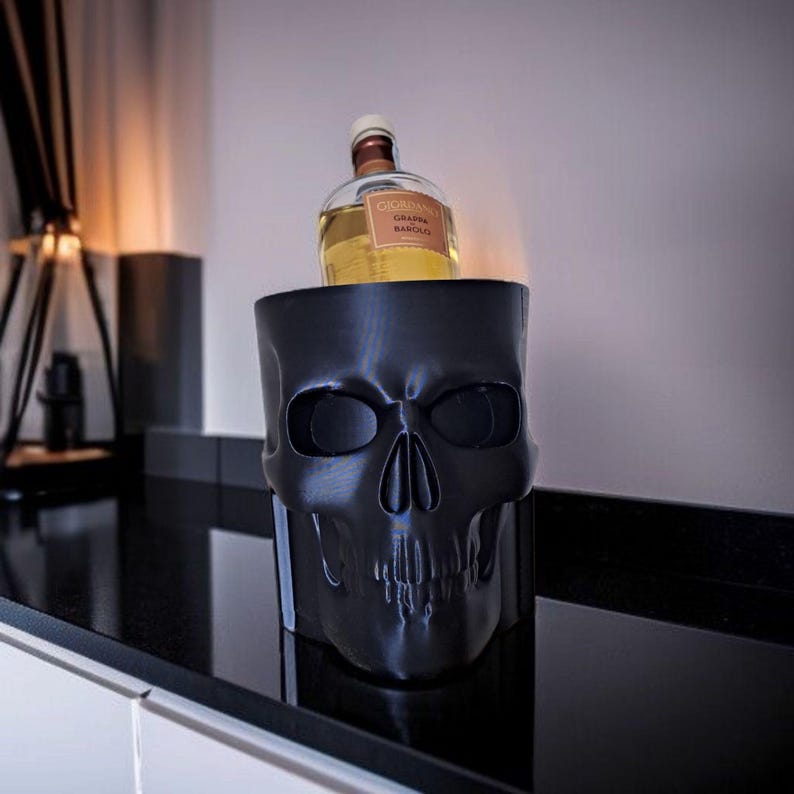May include: A black skull-shaped container holding a bottle of liquor. The container has eye sockets, a nose cavity, and a detailed jawline. The bottle has a tan label and a brown cap. The container sits on a black surface.