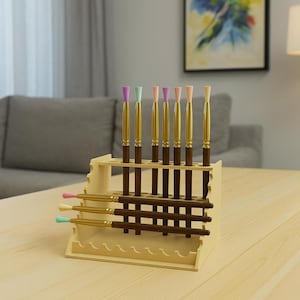 May include: A wooden brush holder with multiple paint brushes. The brushes have gold handles and brown stems, with pastel-colored tips. The holder is light-colored wood and has multiple slots for the brushes.