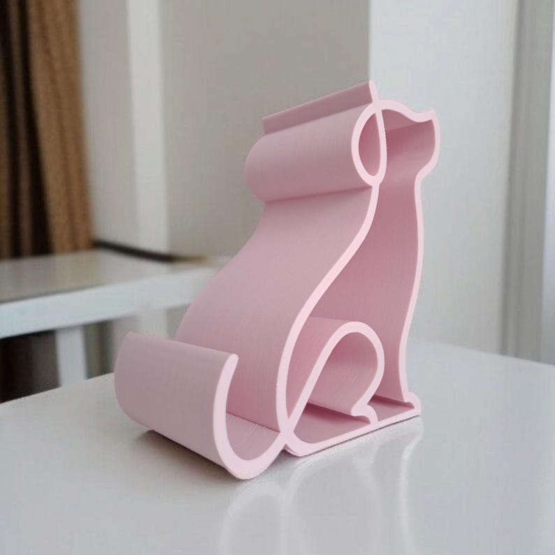 May include: A light pink, stylized cat-shaped phone stand. The stand is made of a solid material and features a continuous line design. The cat is sitting with its tail curled and ears perked up. The stand is on a white surface.