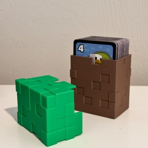 May include: Two cube-shaped card holders. One is bright green, the other is brown and contains a stack of cards. The holders have a pixelated design, with square cutouts. The cards have a blue background and a number 4.