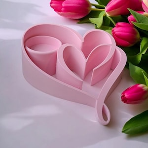 May include: A light pink heart-shaped decorative tray with a cylindrical compartment and two heart-shaped compartments. Pink tulips with green leaves are in the background. The tray is on a white surface.