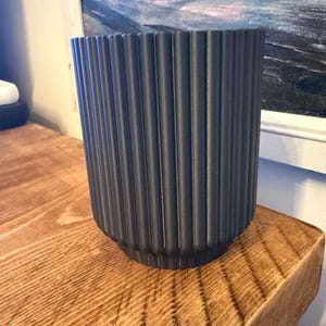 May include: A dark gray, ribbed ceramic planter. The cylindrical pot has vertical grooves and sits on a wooden surface. The planter is empty and ready for a plant. The background includes a painting in a white frame.