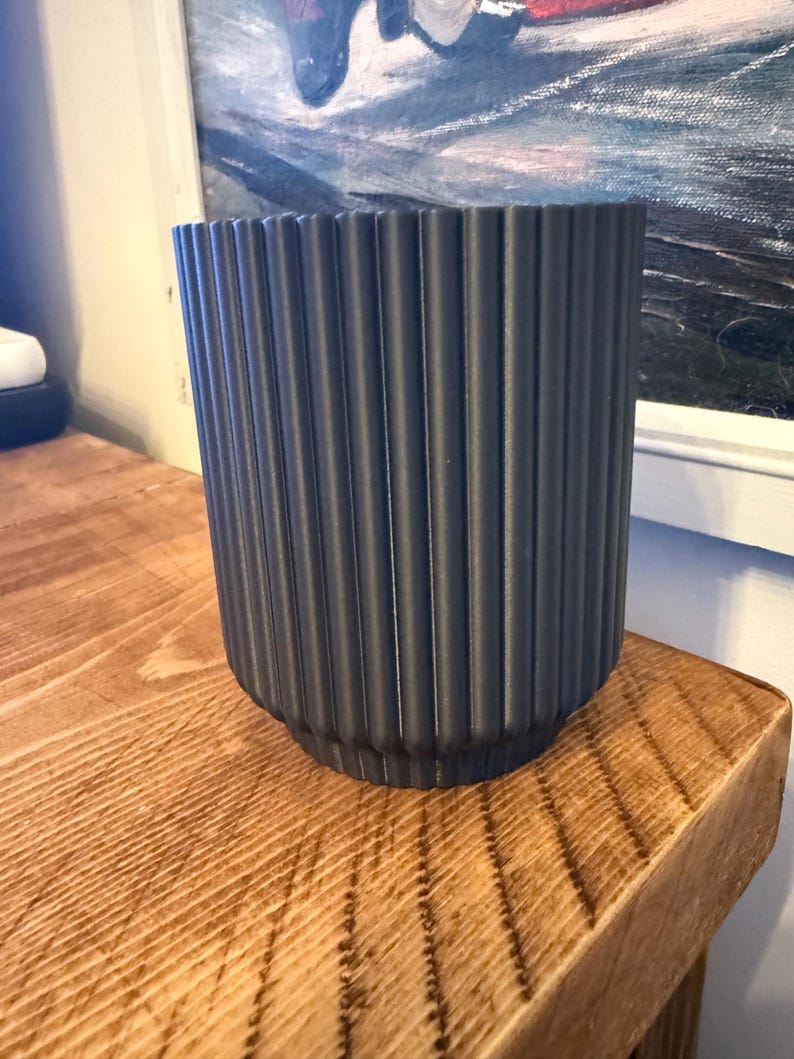 May include: A dark gray, ribbed ceramic planter. The cylindrical pot has vertical grooves and sits on a wooden surface. The planter is empty and ready for a plant. The background includes a painting in a white frame.