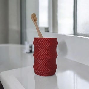 May include: A red toothbrush holder with a zigzag pattern, holding a wooden toothbrush. The holder is cylindrical and sits on a white surface, providing a stylish and functional bathroom accessory.