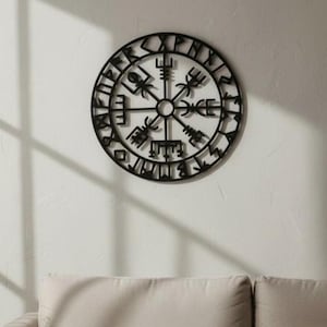 Peut inclure: Une &oelig;uvre d'art murale en m&eacute;tal noir repr&eacute;sentant le Vegv&iacute;sir, une boussole viking. Le design circulaire est mont&eacute; sur un mur blanc au-dessus d'un canap&eacute; beige. Le Vegv&iacute;sir est un symbole de guidage et de protection.