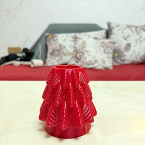 May include: A bright red, decorative tree-shaped object. The tree has a layered design with a series of leaf-like shapes. The object is sitting on a light-coloured surface, with a red background and pillows in the background.