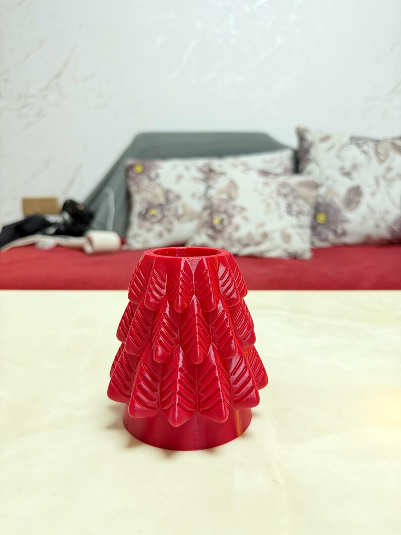 May include: A bright red, decorative tree-shaped object. The tree has a layered design with a series of leaf-like shapes. The object is sitting on a light-coloured surface, with a red background and pillows in the background.