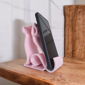 May include: A light pink phone stand shaped like a dog, holding a black smartphone. The stand is made of a smooth material and sits on a wooden surface. The dog design is stylized and modern.