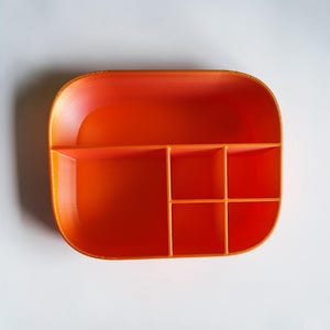 May include: An orange, rectangular organizer with rounded corners and multiple compartments. The organizer is divided into six sections of varying sizes, ideal for storing small items. The bright orange color provides a vibrant and modern aesthetic.