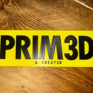 Pu&ograve; includere: Un cartello rettangolare giallo con la scritta "PRIM3D & CREATE3D" in nero, con lettere in rilievo. Il cartello &egrave; posto su una superficie di legno con un motivo a venature visibile. Il design &egrave; semplice e moderno.