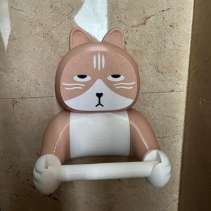 May include: A novelty cat-shaped toilet paper holder in shades of tan, white, and brown. The holder features a cat face with a grumpy expression and holds a white toilet paper roll. The design is whimsical and decorative.
