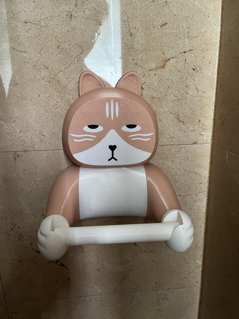 May include: A novelty cat-shaped toilet paper holder in shades of tan, white, and brown. The holder features a cat face with a grumpy expression and holds a white toilet paper roll. The design is whimsical and decorative.