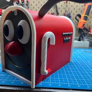 May include: A red and black novelty mailbox with a cartoon face design. The mailbox has a white trim, a red nose, and large white eyes. The text "POSTMAN MAIL" is on the side. A white candy cane handle is on the side.