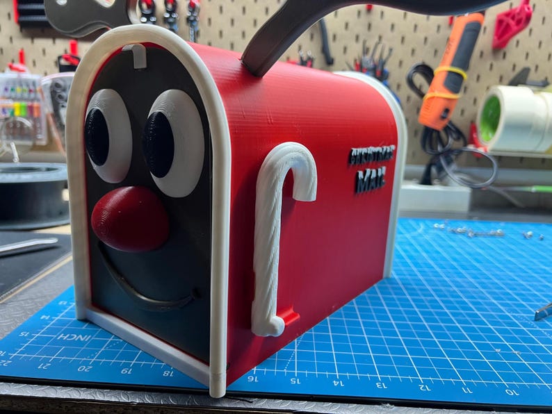 May include: A red and black novelty mailbox with a cartoon face design. The mailbox has a white trim, a red nose, and large white eyes. The text "POSTMAN MAIL" is on the side. A white candy cane handle is on the side.