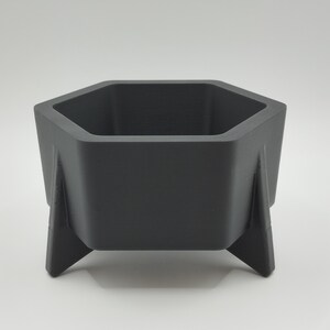May include: A dark gray, hexagonal planter with three angled legs. The planter has a modern, geometric design and is suitable for small plants or succulents. The interior is empty, ready for planting.