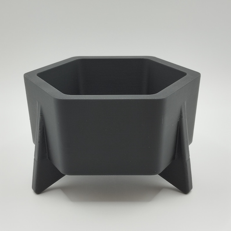 May include: A dark gray, hexagonal planter with three angled legs. The planter has a modern, geometric design and is suitable for small plants or succulents. The interior is empty, ready for planting.