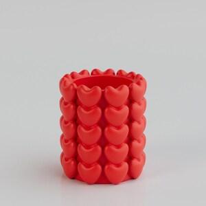 May include: A vibrant red cylindrical container adorned with rows of heart-shaped designs. The container is a solid color and has a smooth texture. The design is repeated around the entire container. This decorative item could be used as a pencil holder or small vase.