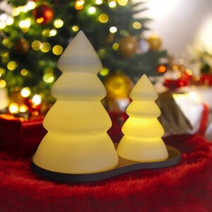 May include: Two white ceramic Christmas tree shaped candle holders with a warm glow. The candle holders are on a black base and are sitting on a red fuzzy surface.