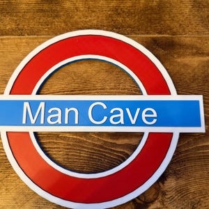May include: A round sign with a red outer ring and a blue center band that reads "Man Cave" in white letters. The sign is set against a wooden background.