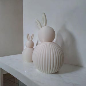 May include: Two off-white ceramic bunny sculptures. One is larger with a ribbed body and the other is smaller with a smooth body. Both have rounded heads and long ears. They are displayed on a white marble surface.