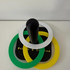 May include: A black handle with a white, green, and yellow ring design. The rings are layered over a black rectangular base with the text "Printed & Created". The object is on a white surface.