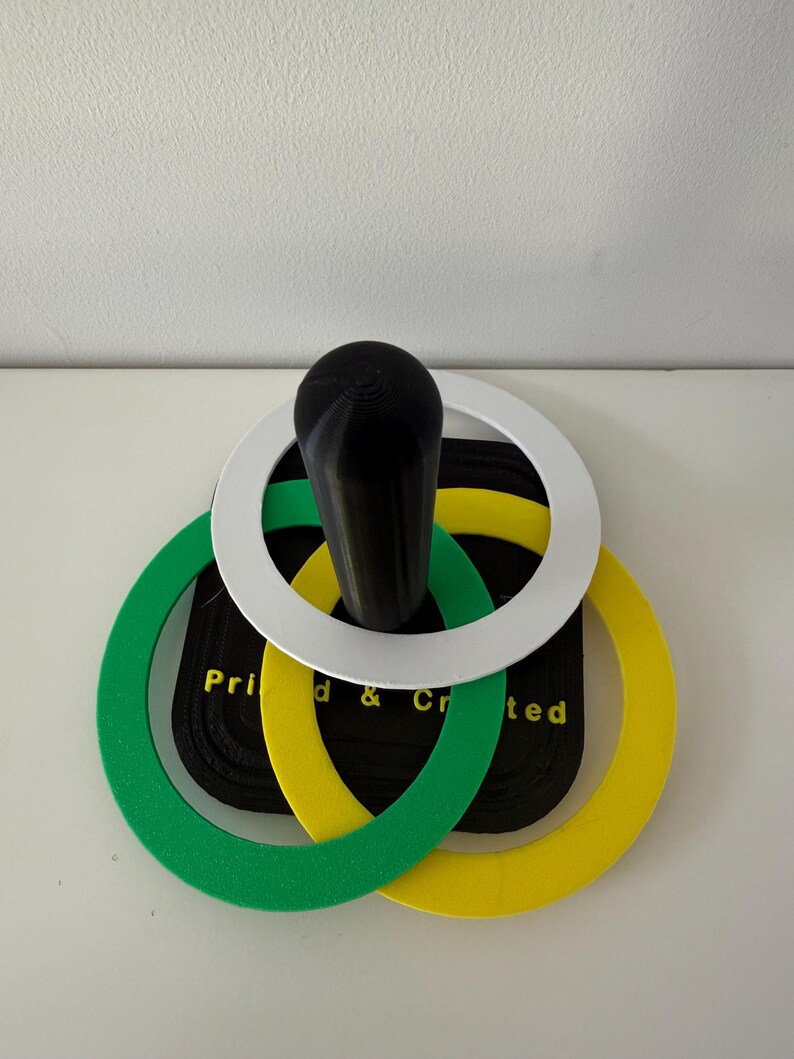 May include: A black handle with a white, green, and yellow ring design. The rings are layered over a black rectangular base with the text "Printed & Created". The object is on a white surface.