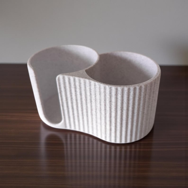 May include: A light gray, textured desk organizer with two circular compartments and a curved slot. The organizer has vertical ribbed detailing and sits on a dark brown surface.