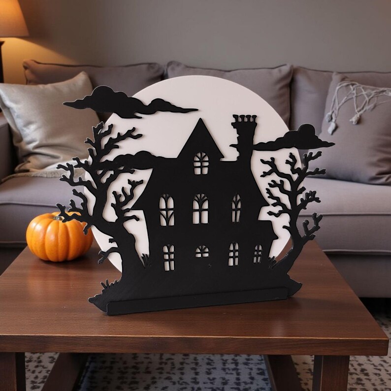 May include: A black silhouette of a haunted house with a white moon backdrop, trees, and clouds. A small orange pumpkin sits to the left. This Halloween decoration is made of wood and is displayed on a brown table.