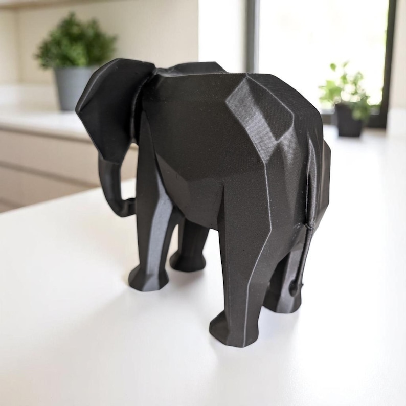 Op de afbeelding: Een zwarte geometrische olifant sculptuur. De sculptuur heeft een facetontwerp, waardoor het een moderne, hoekige uitstraling heeft. De olifant staat op een wit oppervlak, met een vage achtergrond met planten en een raam.