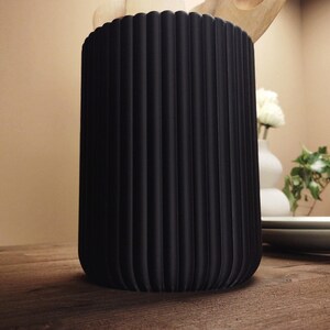 May include: A black, cylindrical container with vertical ridged detailing. The container is on a wooden surface. The background includes a white vase with flowers, plates, and greenery.