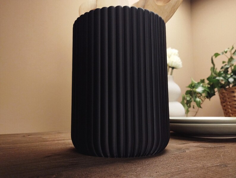May include: A black, cylindrical container with vertical ridged detailing. The container is on a wooden surface. The background includes a white vase with flowers, plates, and greenery.