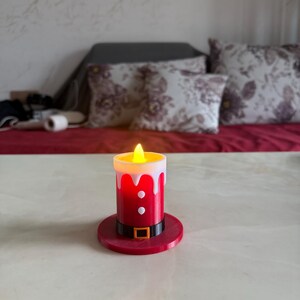 May include: A red and white LED candle with a flickering yellow light, designed to resemble Santa Claus. The candle has a black belt with a gold buckle and sits on a matching red base. A festive decorative item.