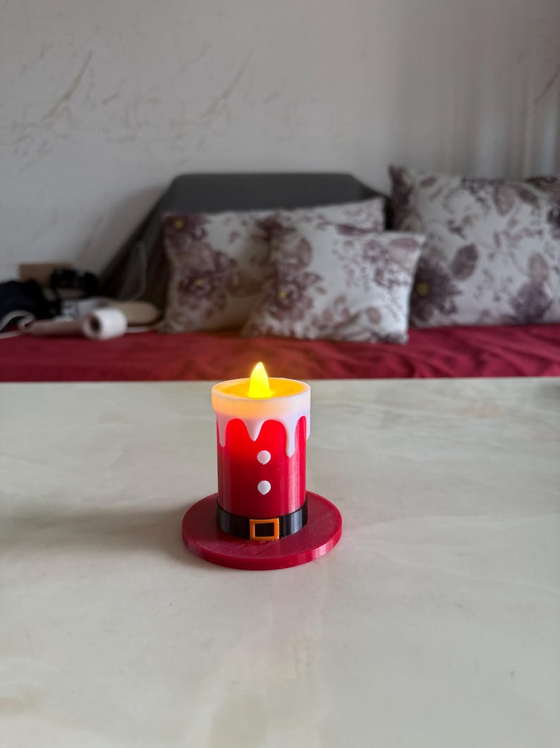 May include: A red and white LED candle with a flickering yellow light, designed to resemble Santa Claus. The candle has a black belt with a gold buckle and sits on a matching red base. A festive decorative item.